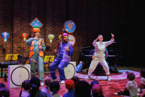 Peformers on a stage with children watching, one is sat on a fake boke made of boxes and two are wearing high-vis jackets and helmets