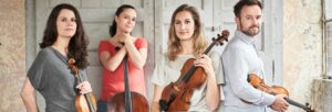 Three women and a man stood in front of a pale wall holding stringed instruments.