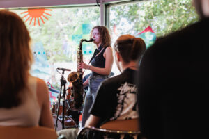 A woman playing a saxophone from the perspective of the crowd, with other crowd members in the foreground