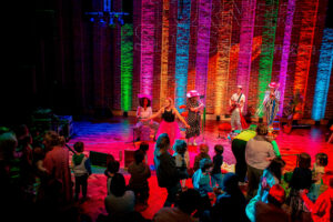 A band in patterned outfits performing on a stage with rainbow lighting behind them.