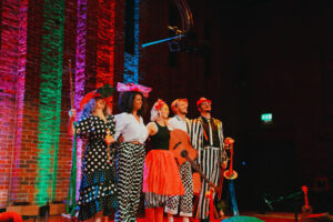 A band wearing black, white and red patterned outfits, standing with their arms around each other on a stage