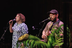 A man and a woman with mics to their mouths, the man is playing a guitar