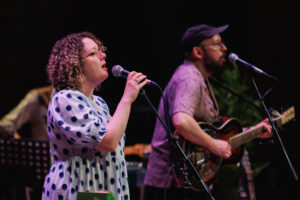 A man and a woman singing into mics, the man is playing a guitar