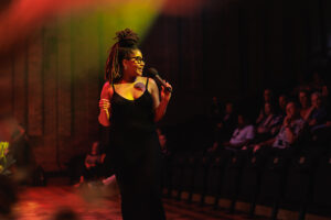 Woman under stage lights in a black dress singing into a microphone and looking towards an audience