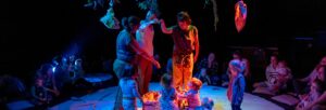 Three women stand under a hanging canopy on a circular stage, surrounded by small children. Parents and children surround the circle.