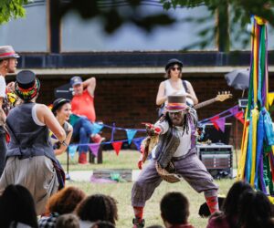 Man with a top hat, colourful waistcoat, checked trousers and colourful socks reaching out to an audience of families with musicians and a maypole in the background