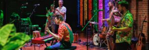 man in dungarees and a bright red shirt playing a keyboard with a band in the background and rainbow coloured lights lighting the brick wall at the back of Turner Sims