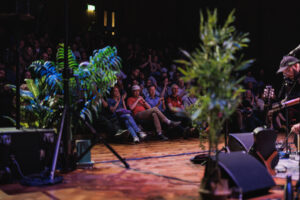 View across stage to audience sitting in seats applauding with big plants and sound equipment in the foreground