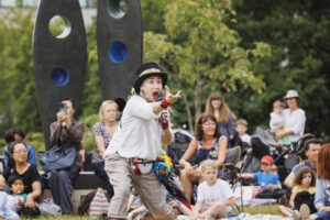 Female performer reaching out in front of her with a microphone in her other hand and a crowd of families in the background.