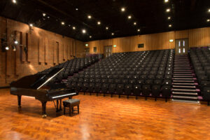 Grand piano under stage lights with black raked seating in the background.