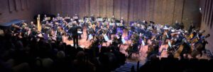 A large orchestra seated on a stage, under lights