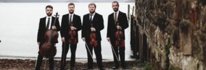 Four men in suits holding stringed instruments and standing on a beach