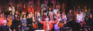 A choir standing on stage. Their director stands at the front, a man wearing a red jacket holding his arms in the air