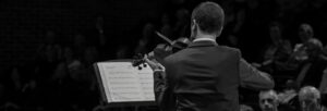 A man stands with his back to the camera playing the violin to an audience. The image is in black and white