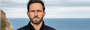 A man with a beard and dark jumper standing in front of the sea