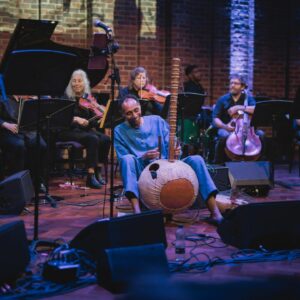 Man sat on the floor of Turner Sims' stage holding a Kora and smiling with string players and a drummer in the background