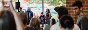 A vocalist and a saxophonist perform in front of a large window as audience members watch and take pictures.