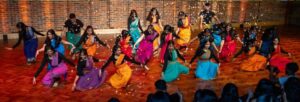 A group of young women wearing brightly coloured saris dancing on stage in front of an audience