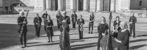 A black and white image of an orchestra with their instruments standing outside a large, pillared building