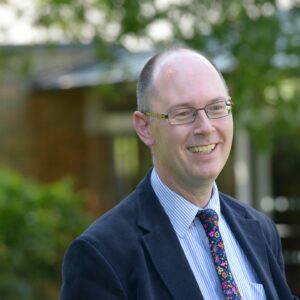 A smiling man wearing glasses and a suit and tie, standing outside with trees in the background